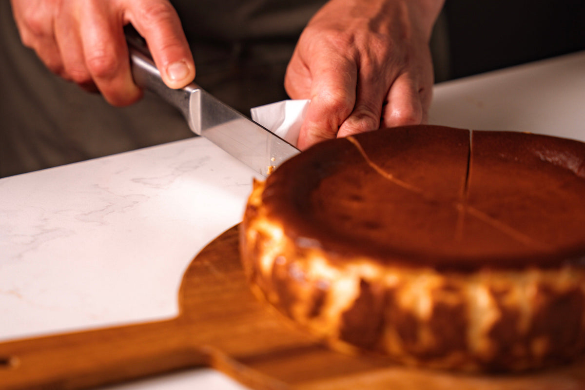 A close-up lifestyle shot of a person's hands slicing a freshly baked Basque burnt cheesecake on a wooden board with a knife.